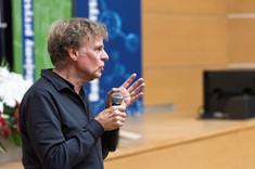 Mężczyzna ubrany na czarno podczas prowadzenia wykładu. W tle zielono-biało-niebieskie banery i drewnopodobna ściana sali wykładowej. Prof. arch. W. Maas, MVRDV.