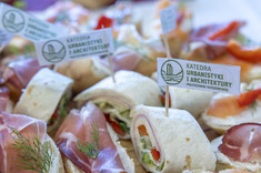 Zbliżenie na poczęstunek. Pośród jedzenia umieszone białe znaczniki z zielonym napisem: Katedra Urbanistyki i Architektury.