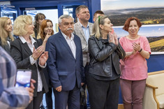 Ujęcie pomieszczenia w którym stoi grupa kilkunastu osób. Wszyscy zwróceni w jednym kierunku, biją brawo i uśmiechają się. W tle na granatowej ścianie zawieszone plansze z dużymi kolorowymi fotografiami.
