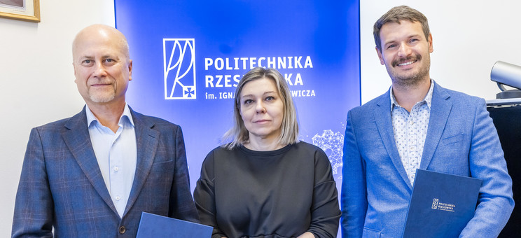 Na tle białej ściany z niebieskim pasem z napisem politechnika rzeszowska stoją obok siebie trzy osoby dwóch mężczyzn i kobieta po środku. Wszyscy elegancko ubrani w różnych odcieniach niebieskiego. Uśmiechają się. Mężczyźni trzymają w rękach okładki z dokumentami.