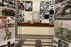 Pomiędzy dwoma ścianami z siatki, z zawieszonymi planszami z kolorowymi mapami miast, widok na ścianę z modelami. Modele na ścianie przeważnie w biało-czarnej kolorystyce.