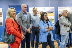 Ujęcie pomieszczenia w którym stoi grupa kilku osób. Wszyscy zwróceni w jednym kierunku, uśmiechają się. W tle na granatowej ścianie zawieszone plansze z dużymi kolorowymi fotografiami.