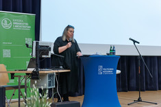 Kobieta ubrana na czarno stoi za pulpitem i przemawia do zebranych. W tle duży biały ekran i zielony baner informacyjny z białym napisem - Katedra Urbanistyki i Architektury. Kierowniczka KUA, prof. arch. A. Sikora.