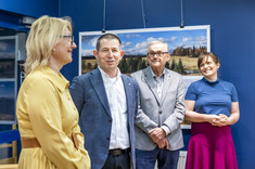 W pomieszczeniu stoją obok siebie cztery osoby, dwóch mężczyzn pośrodku i po bokach kobiety. Rozmawiają ze sobą i uśmiechają się. W tle na granatowej ścianie zawieszone plansze z dużymi kolorowymi fotografiami.