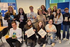 W pomieszczeniu zgromadzonych jest razem kilkanaście osób. Jedni stoją, drudzy przysiadają, wszyscy pozują razem do zdjęcia i trzymają w rękach różne prace plastyczne. Wszyscy się uśmiechają.