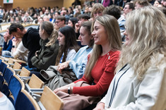 Zbliżenie na grupę studentów słuchających wykładu. W tle sala wykładowa wypełniona w całości słuchaczami wykładu. Na pierwszym planie studentki kierunku architektura.