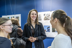 Trzy młode kobiety podczas rozmowy. W tle plansze ze zdjęciami. Na zdjęciu studentki kierunku architektura.