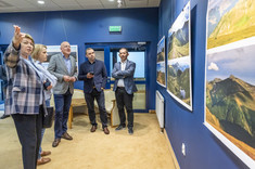 W pomieszczeniu kilka osób stoi przed prezentowanymi na ścianie fotografiami. Kobieta na pierwszym planie wskazuje coś ręka pozostałym rozmówcom.