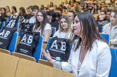 Małgorzata Tomczak siedząc w sali wykładowej zadeje pytanie prelegentowi. W tle jej postaci widać innych słuchaczy wykładu oraz pakiety wydarzeniowe od sponsorów.