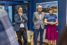 W pomieszczeniu stoją obok siebie trzy osoby, dwóch mężczyzn i kobieta po lewej stronie. Wszyscy zwróceni w jednym kierunku. W tle na granatowej ścianie zawieszone plansze z dużymi kolorowymi fotografiami.