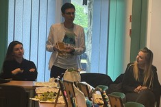 W pomieszczeniu na długim stole wystawione różne prace plastyczne. Za stołem stoi i tłumaczy coś mężczyzna, słuchają go dwie kobiety siedzące za stołem.