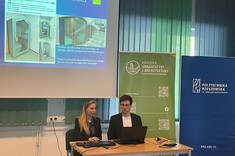 W lekko przyciemnionym pomieszczeniu po środku wysoko zawieszony jasny ekran, przed nim przy stole siedzi dwóch prelegentów, mężczyzna i kobieta. W tle kilka wysokich na około 2 metry plansz w zielonym lub niebieskim kolorze z białymi napisami.