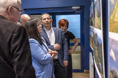 Kilka osób zgromadzonych przy planszach z fotografiami zawieszonymi na ciemnej ścianie. Przyglądają się uważnie szczegółom zdjęć.