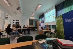 W lekko przyciemnionym pomieszczeniu widocznych jest po lewej stronie kilka osób słuchających wykładu. Po prawej stronie wysoko zawieszony jasny ekran, przed nim siedzi przy stole prelegentka.