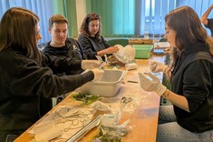 W pomieszczeniu przy długim stole siedzą cztery osoby, trzy kobiety i jeden mężczyzna. Wszyscy ubrani na ciemno. Na stole rozłożone różne materiały, zgromadzeni wykonują z nich prace plastyczne.