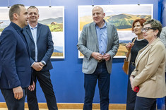 W pomieszczeniu stoi obok siebie pięć osób, trzech mężczyzn po lewej stronie i dwie kobiety po prawej. Rozmawiają ze sobą i uśmiechają się. W tle na granatowej ścianie zawieszone plansze z dużymi kolorowymi fotografiami.