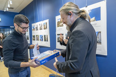 Mężczyzna w ciemnej marynarce wręcza nagrodę mężczyźnie w ciemnym golfie. W tle plansze ze zdjęciami. Na zdjęciu: arch. J. Dudek i arch. W. Matłok.