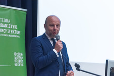  Mężczyzna w garniturze przemawia do zebranych. W tle biały ekran i zielony baner. Prezydent Miasta Rzeszowa K. Fijołek.