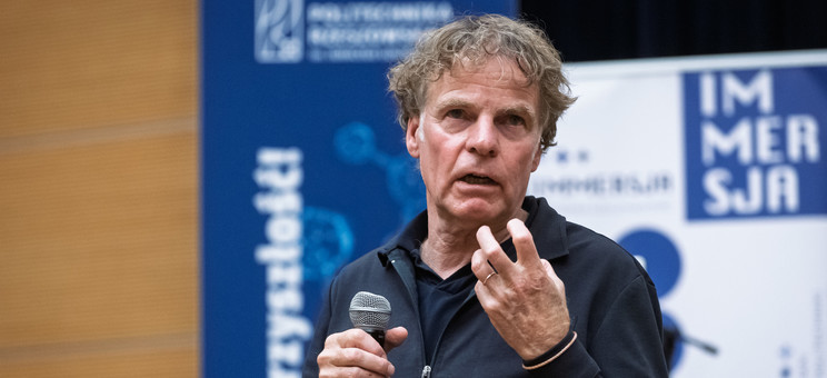 Mężczyzna, prelegent ubrany na czarno, podczas prowadzenia wykładu. W tle biało-niebieskie banery informacyjne. Prof. arch. Winy Maas, MVRDV