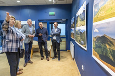 W pomieszczeniu kilka osób stoi przed prezentowanymi na ścianie fotografiami. Kobieta na pierwszym planie wskazuje coś ręka pozostałym rozmówcom.