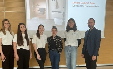 Na tle ekranu z wyświetloną prezentacją grupa sześciu osób. Po lewej mężczyzna w ciemnym ubraniu. Pośrodku grupy kobiet w białych podkoszulkach prelegentka w ciemnym ubraniu. Wszyscy się uśmiechają.