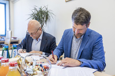 Dwóch mężczyzn elegancko ubranych siedzi przy stole i podpisuje dokumenty.