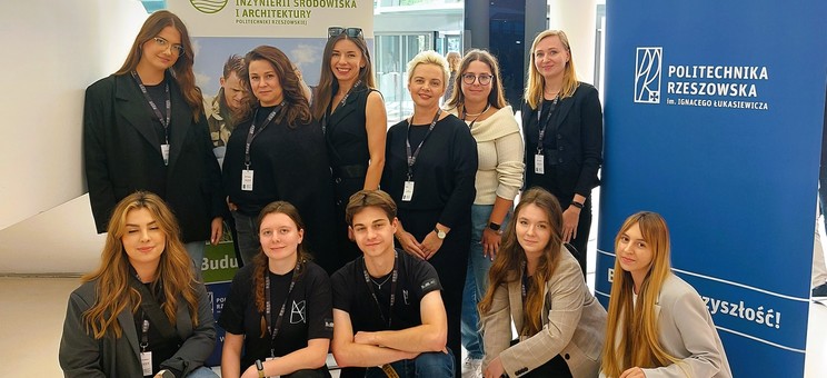 Na zdjęciu grupa 11 osób – 5 kobiet, pracowników Zakładu Urbanistyki i Architektury oraz pięć studentek i student Koła Naukowego Architektury Miejsc Społecznych na tle biało-niebieskiej planszy z napisem Politechniki Rzeszowskiej i jasnej planszy Wydziału BIŚiA w nowoczesnym budynku Centrum Kongresowego.