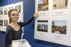 Zbliżenie na młoda kobietę z jasnymi włosami i w okularach. Kobieta wskazuje ręką na zdjęcie, w drugiej ręce trzyma nagrodę. Na zdjęciu: stud. arch. Z. Dąbrowska.
