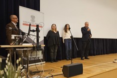 Na scenie znajduje się cztery osoby. Stoją na scenie w audytorium. Po lewej stronie stoi sprzęt komputerowy. W tle znajduje się duży ekran projekcyjny. Scena wykonana jest z jasnego drewna, a w tle widzimy ciemne kotary.