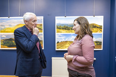 Na pierwszym planie w pomieszczeniu stoją dwie osoby, kobieta w różowej bluzce i mężczyzna w ciemnej marynarce. Są zwróceni do siebie i rozmawiają ze sobą. W tle na granatowej ścianie zawieszone plansze z dużymi kolorowymi fotografiami.