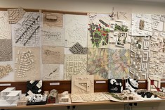 Zbliżenie na ścianę pokrytą modelami miast. Modele w jasnych kolorach. Nieliczne z akcentami kolorystycznymi. Wzdłuż ściany stoją stoły z poustawianymi na nich modelami budynków.