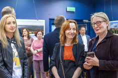 Na pierwszym planie trzy kobiety stojące obok siebie, uśmiechają się do fotografii.  W tle kilkanaście osób na tle ciemnej ściany z prezentowanymi zdjęciami.