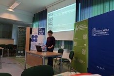 W lekko przyciemnionym pomieszczeniu po środku wysoko zawieszony jasny ekran, przed nim przy stole stoi prelegentka. W tle kilka wysokich na około 2 metry plansz w zielonym lub niebieskim kolorze z białymi napisami.