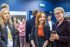 Na pierwszym planie trzy kobiety stojące obok siebie, uśmiechają się do fotografii.  W tle kilkanaście osób na tle ciemnej ściany z prezentowanymi zdjęciami.