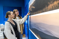 Przed planszami z fotografiami pejzaży górskich stoją dwie kobiety. Uśmiechają się. Jedna z nich wskazuje drugiej jakiś fragment zdjęcia.