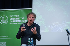 Mężczyzna ubrany na czarno prowadzi wykład. W tle jasny ekran prezentacji i w rogu zielony baner. Prof. arch. W. Maas, MVRDV.