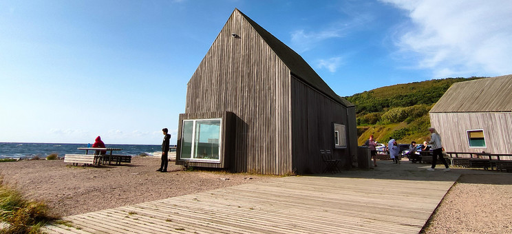 Zdjęcie: Pośrodku rozległej przestrzeni w naturalnym surowym krajobrazie zlokalizowany parterowy budynek. Bryła prosta z wysokim dachem, całość wykończona pionowym szarym deskowaniem. W tle zielone wzgórze i jaskrawo niebieskie niebo.