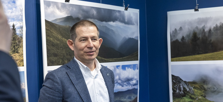 Na pierwszym planie stoi autor wystawy, mężczyzna w ciemnej marynarce.  W tle na granatowej ścianie zawieszone plansze z dużymi kolorowymi fotografiami pejzaży górskich.
