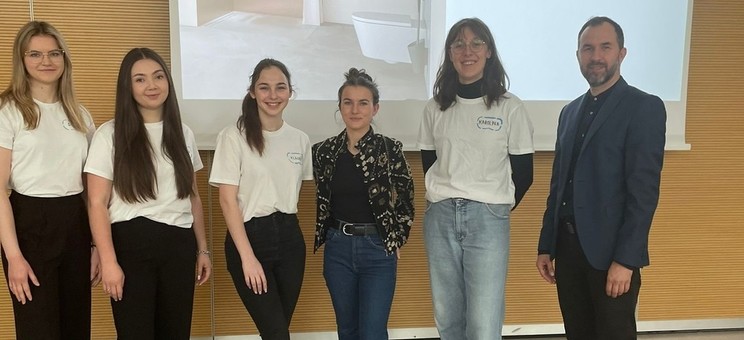 Na tle ekranu z wyświetloną prezentacją grupa sześciu osób. Po lewej mężczyzna w ciemnym ubraniu. Pośrodku grupy kobiet w białych podkoszulkach prelegentka w ciemnym ubraniu. Wszyscy się uśmiechają.