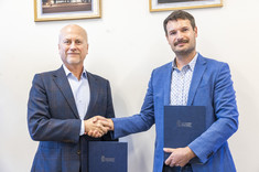 Dwóch mężczyzn elegancko ubranych stoi na tle białej ściany. W rękach trzymają okładki z dokumentami. Podają sobie ręce i uśmiechają się.