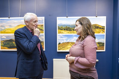 Na pierwszym planie w pomieszczeniu stoją dwie osoby, kobieta w różowej bluzce i mężczyzna w ciemnej marynarce. Są zwróceni do siebie i rozmawiają ze sobą. W tle na granatowej ścianie zawieszone plansze z dużymi kolorowymi fotografiami.