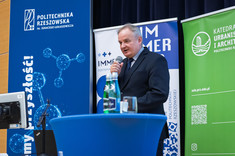 Mężczyzna w ciemnym garniturze stoi za niebieskim pulpitem, przemawia do zebranych. W tle biało-niebieskie i zielono-białe banery informacyjne jednostek politechniki. Dziekan WBIŚiA, prof. L. Lichołai.