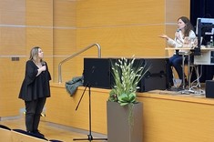 Zdjęcie przedstawia moment dyskusji w nowoczesnej sali audytoryjnej. Po lewej stronie kadru stoi kobieta ubrana na czarno z mikrofonem w dłoni. Po prawej stronie siedzi prelegentka z mikrofonem, odpowiadając na pytanie.