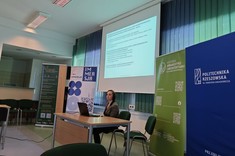 W lekko przyciemnionym pomieszczeniu po środku wysoko zawieszony jasny ekran, przed nim przy stole siedzi prelegentka. W tle kilka wysokich na około 2 metry plansz w zielonym lub niebieskim kolorze z białymi napisami.