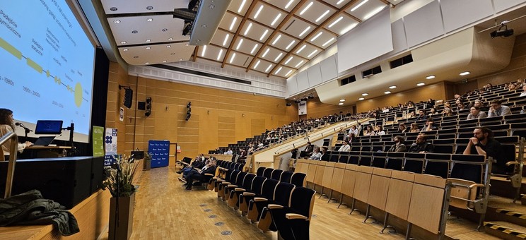 Zdjęcie przedstawia dużą aulę wykładową podczas trwania prezentacji. Po lewej stronie kadru widoczna jest prelegentka — kobieta z długimi, kręconymi włosami — stojąca za pulpitem z laptopem i mikrofonem. Na ekranie za nią widnieje slajd. Widownia zajmuje większość kadru — sala jest wypełniona liczną grupą uczestników, którzy z zainteresowaniem słuchają wystąpienia. 