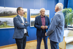 Trzech mężczyzn, ubranych w różnych odcieniach niebieskiego garniturach stoi obok siebie i rozmawia. Uśmiechają się do siebie. W tle na granatowej ścianie zawieszone plansze z dużymi kolorowymi fotografiami.