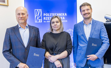 Na tle białej ściany z niebieskim pasem z napisem politechnika rzeszowska stoją obok siebie trzy osoby dwóch mężczyzn i kobieta po środku. Wszyscy elegancko ubrani w różnych odcieniach niebieskiego. Uśmiechają się. Mężczyźni trzymają w rękach okładki z dokumentami.