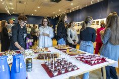 Wnętrze z ciemno niebieskimi ścianami. We wnętrzu, w kilku luźnych grupach, zgromadzone osoby odwiedzjące wystawę. Na pierwszym planie stół z poczęstunkiem.