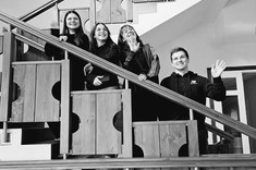Zdjęcie czarno białe. Na schodach za drewnianą balustradą stoi na różnych wysokościach czwórka młodych osób, trzy kobiety i jeden mężczyzna. Wszyscy spoglądają prosto w obiektyw i uśmiechają się.