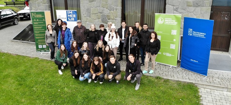 Zdjęcie wykonane z wysokości kilku metrów. Grupa około dwudziestu osób ubranych na sportowo pozuje do zdjęcia. Wszyscy spoglądają do góry. W tle kamienna ściana budynku.